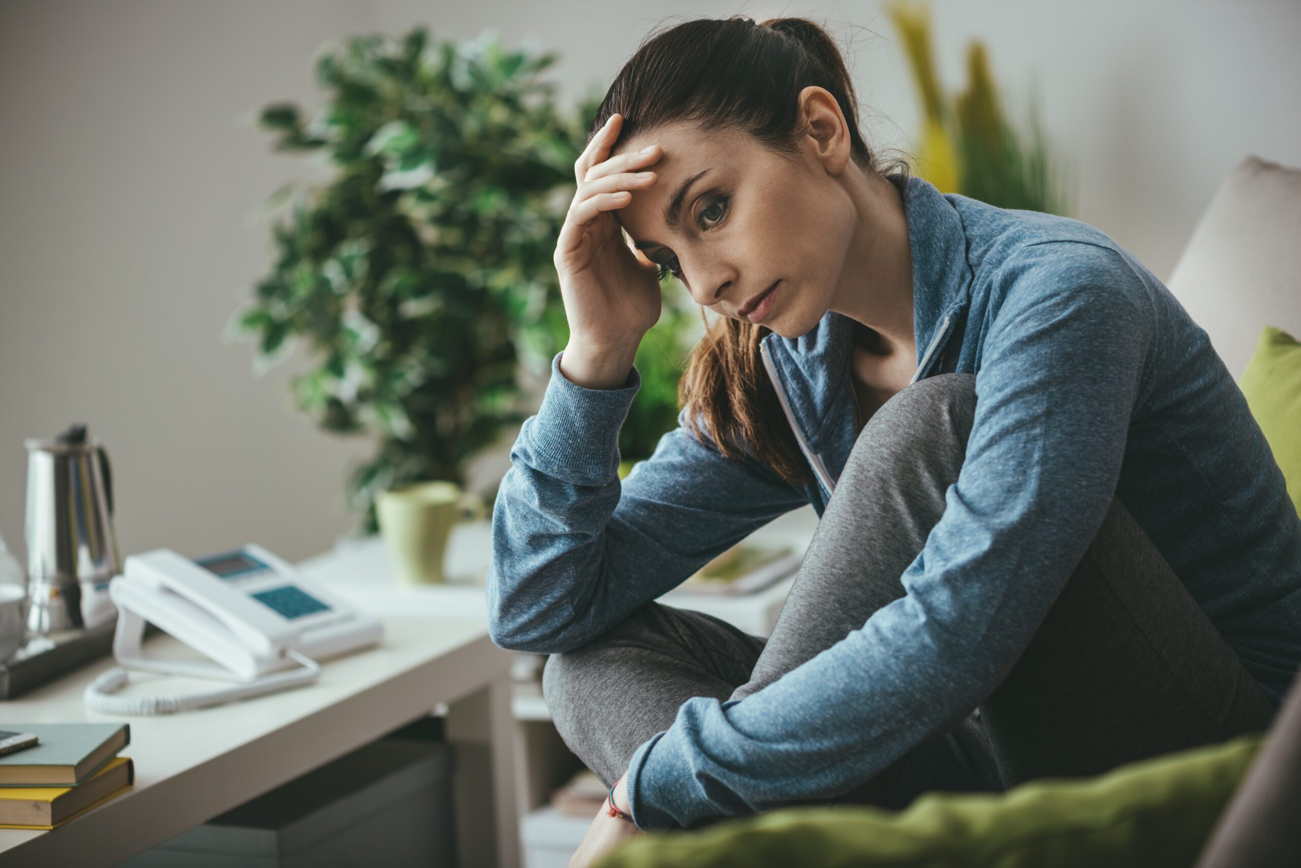 Sad depressed woman at home sitting on the couch, looking down and touching her forehead, loneliness and pain concept