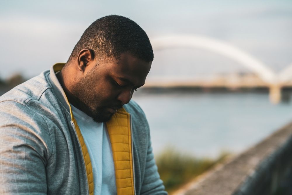 Man standing outdoors near water with his head down, appearing deep in thought or emotional