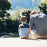 Father and young son sitting side by side on a patio, seen from behind, sharing a quiet moment together in a sunny backyard.