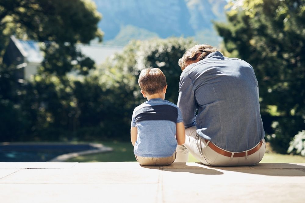 Father and young son sitting side by side on a patio, seen from behind, sharing a quiet moment together in a sunny backyard.