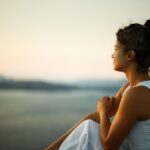 Woman sitting outdoors near water at sunset, gazing thoughtfully into the distance with her arms resting on her knees, wearing a white sleeveless top.