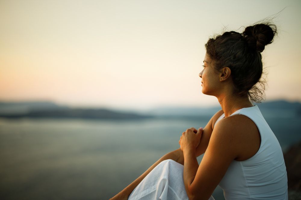 Woman sitting outdoors near water at sunset, gazing thoughtfully into the distance with her arms resting on her knees, wearing a white sleeveless top.
