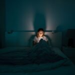 A woman sitting up in bed in a dark room, illuminated only by the glow of her smartphone screen as she looks at it, suggesting late-night phone use or doomscrolling.