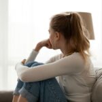 A young woman sits on a couch with her knees pulled up to her chest, resting her chin on her hand. She looks thoughtfully out a bright window, appearing pensive or worried.