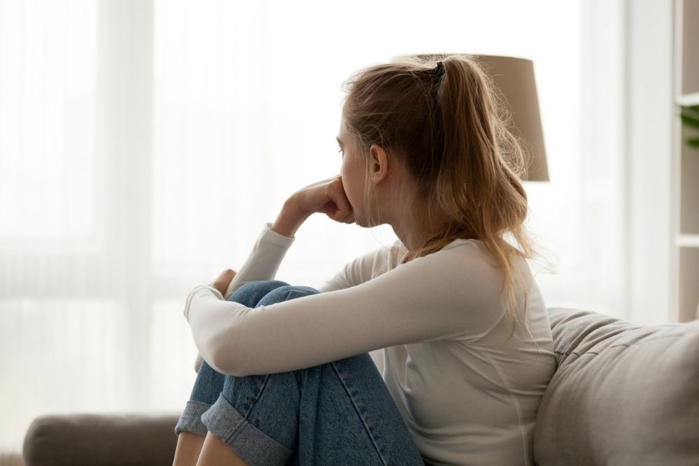 A young woman sits on a couch with her knees pulled up to her chest, resting her chin on her hand. She looks thoughtfully out a bright window, appearing pensive or worried.