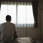 A man sits on the edge of a bed in a dimly lit room, facing a window with closed curtains, suggesting a mood of loneliness, reflection, or depression.