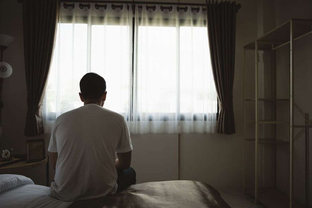 A man sits on the edge of a bed in a dimly lit room, facing a window with closed curtains, suggesting a mood of loneliness, reflection, or depression.