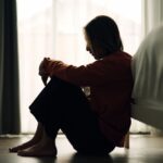 Silhouette of a woman sitting on the floor by her bed with knees pulled to her chest, head bowed, appearing deep in thought or emotionally distressed.