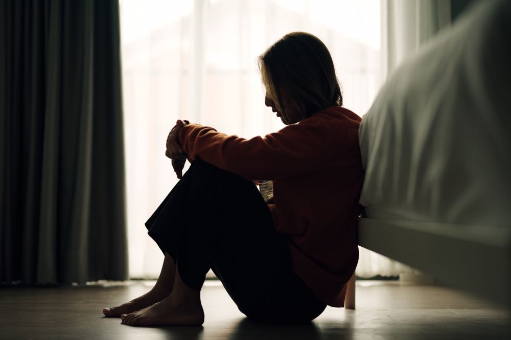 Silhouette of a woman sitting on the floor by her bed with knees pulled to her chest, head bowed, appearing deep in thought or emotionally distressed.