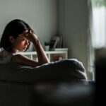 A woman sits on a couch with her head resting on her hand, appearing stressed or overwhelmed, in a dimly lit living room.