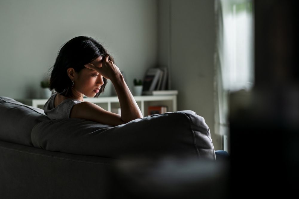 A woman sits on a couch with her head resting on her hand, appearing stressed or overwhelmed, in a dimly lit living room.