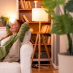 Woman relaxing on a white couch with a peaceful smile, arms behind her head, in a cozy, softly lit living room with bookshelves and houseplants.