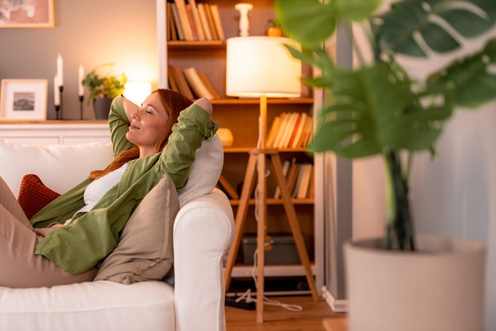 Woman relaxing on a white couch with a peaceful smile, arms behind her head, in a cozy, softly lit living room with bookshelves and houseplants.