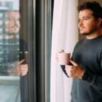 Man holding a coffee mug while standing by a window and looking outside thoughtfully.