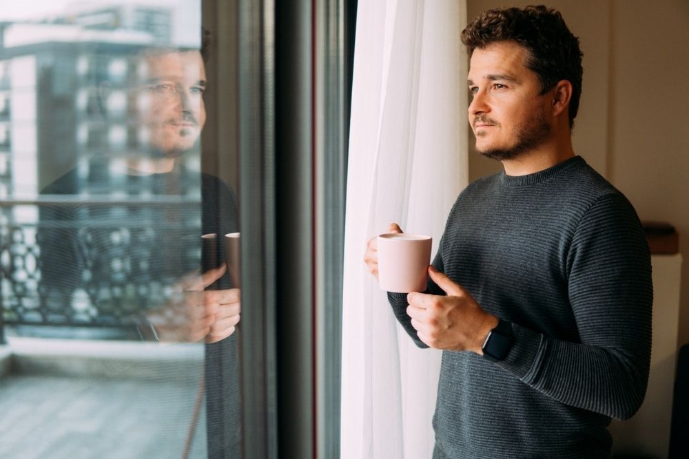 Man holding a coffee mug while standing by a window and looking outside thoughtfully.
