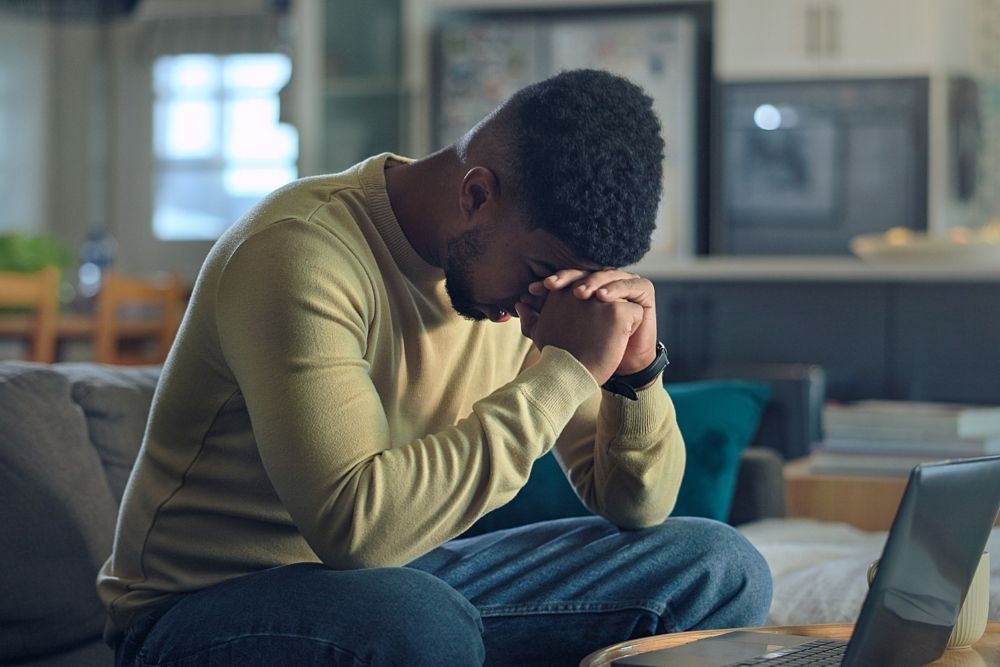 Man sitting on a couch with his head bowed and hands clasped in front of his face, appearing stressed or overwhelmed, with a laptop nearby.