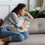A woman sits curled up on a couch, hugging a pillow and looking distressed, with her hand on her forehead in thought.