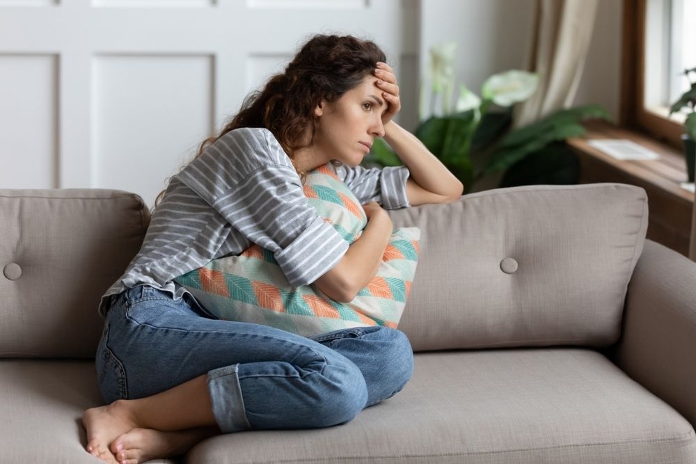 A woman sits curled up on a couch, hugging a pillow and looking distressed, with her hand on her forehead in thought.