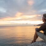 Man sitting on a rock overlooking the ocean at sunset, appearing calm and reflective as he gazes at the horizon.