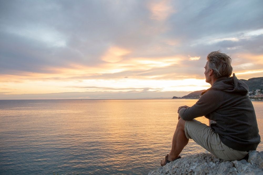 Man sitting on a rock overlooking the ocean at sunset, appearing calm and reflective as he gazes at the horizon.
