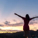 A woman standing outdoors at sunrise with her arms outstretched, embracing the open sky and peaceful landscape below.