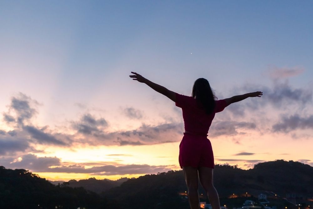 A woman standing outdoors at sunrise with her arms outstretched, embracing the open sky and peaceful landscape below.
