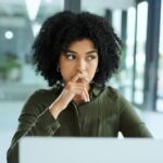 A woman with curly hair sits at a laptop, resting her hand near her mouth with a thoughtful, concerned expression in a bright office setting.