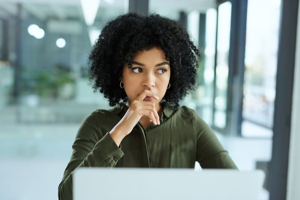 A woman with curly hair sits at a laptop, resting her hand near her mouth with a thoughtful, concerned expression in a bright office setting.
