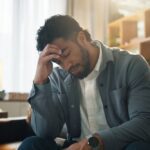 A man sitting indoors with his head bowed and hand on his forehead, appearing stressed, overwhelmed, or deep in thought.