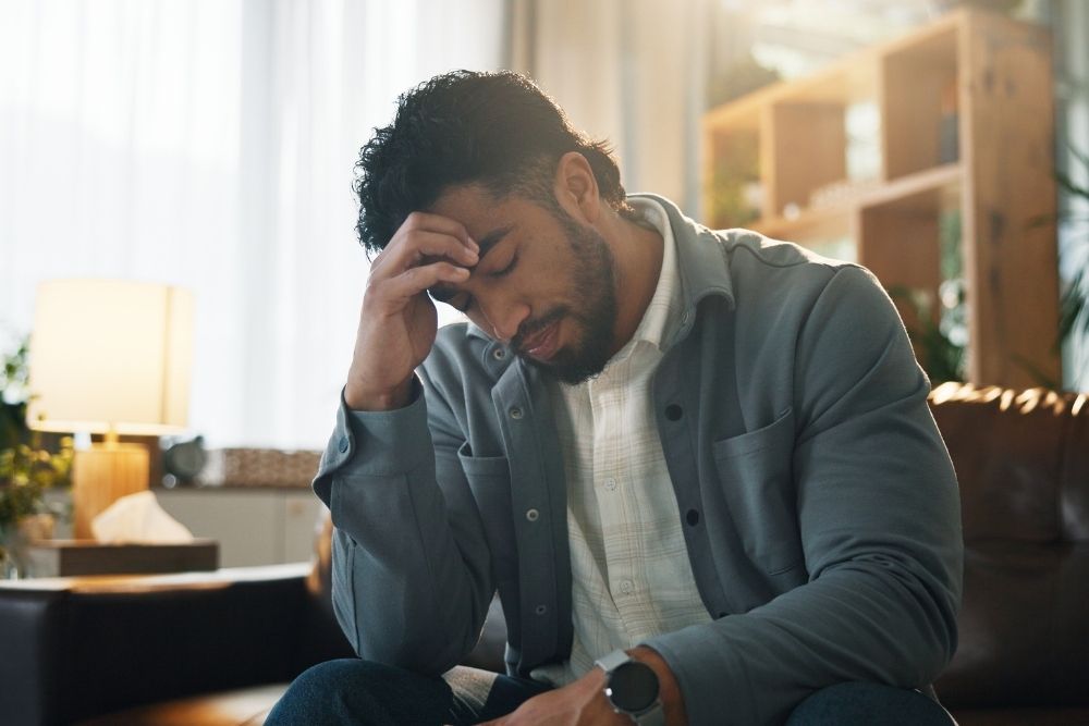 A man sitting indoors with his head bowed and hand on his forehead, appearing stressed, overwhelmed, or deep in thought.