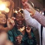 A group of friends smile and raise sparklers together during an indoor New Year’s celebration.