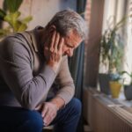 Older adult sitting by a window with head resting in hand, appearing distressed or deep in thought in a quiet indoor space.