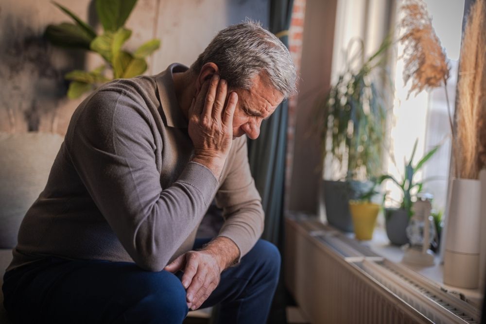 Older adult sitting by a window with head resting in hand, appearing distressed or deep in thought in a quiet indoor space.