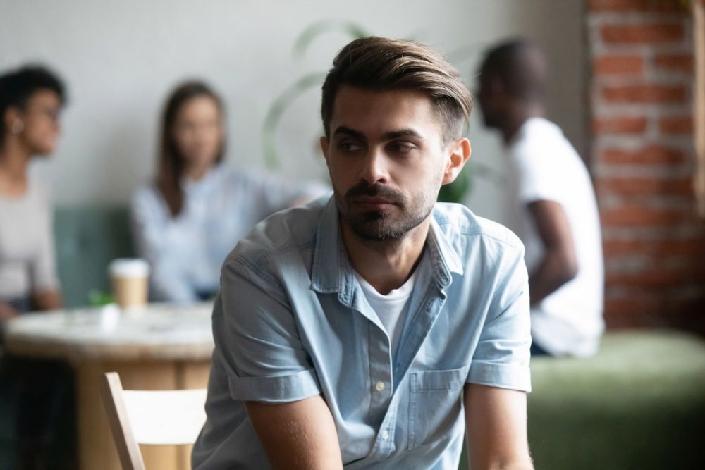 Eagle View - Why Neurodivergent Individuals Often Struggle With Mental Health Challenges - Edited. A man sits looking upset and dejected as friends in the foreground talk amongst themselves about