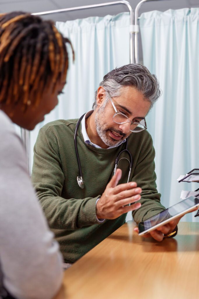 A male doctor holds a tablet computer while reviewing test results with a black female patient. They are seated next to each other in a medical office.