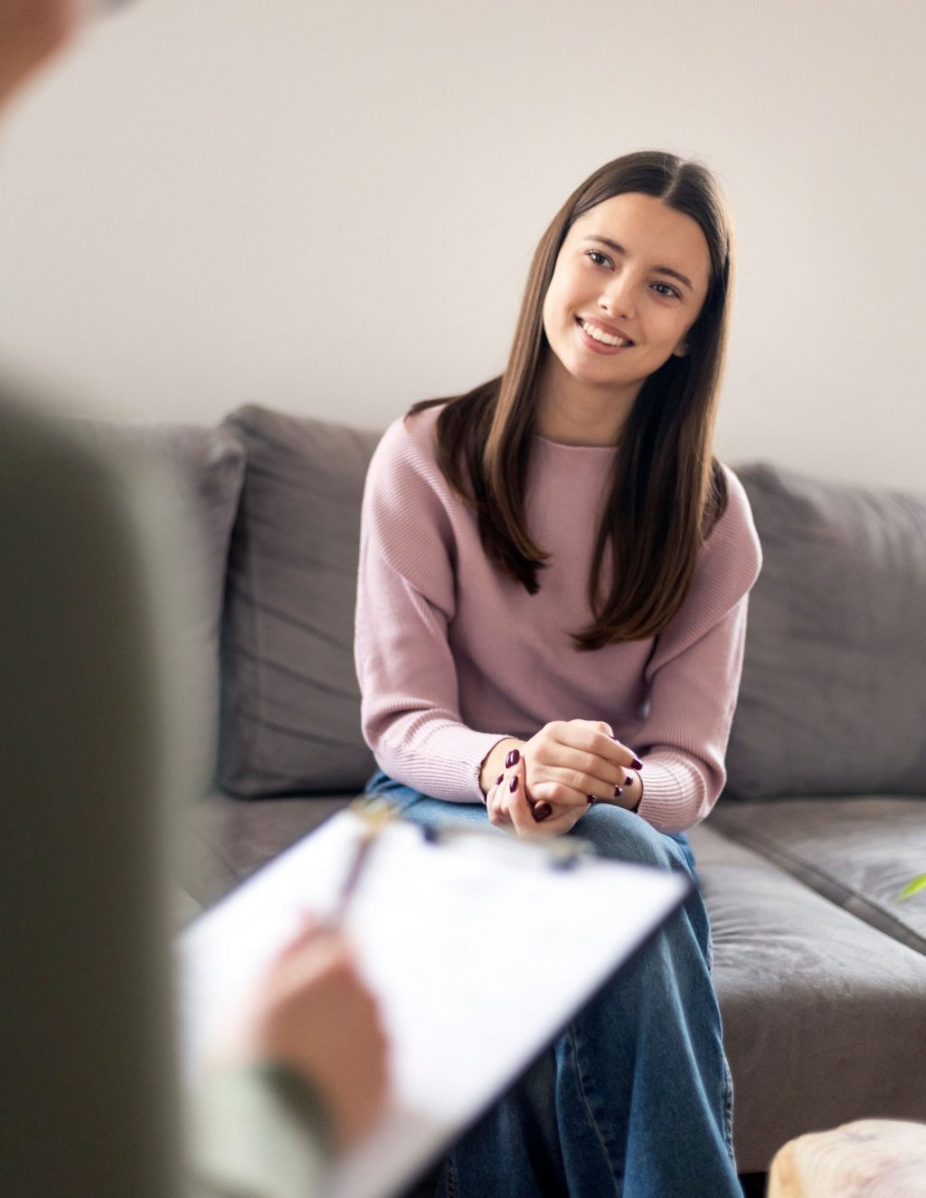 Teenage girl sitting on a grey couch talking to a psychiatrist in a comfortable clinical setting.